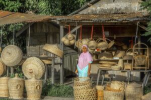 Woman standing amidst hand-woven baskets outside a rustic workshop in Aceh, Indonesia.