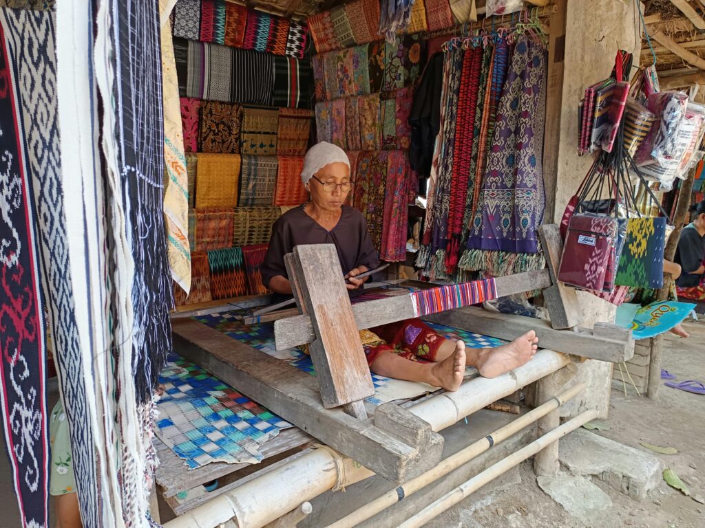 Elderly woman weaving traditional fabric in West Nusa Tenggara, Indonesia market stall.