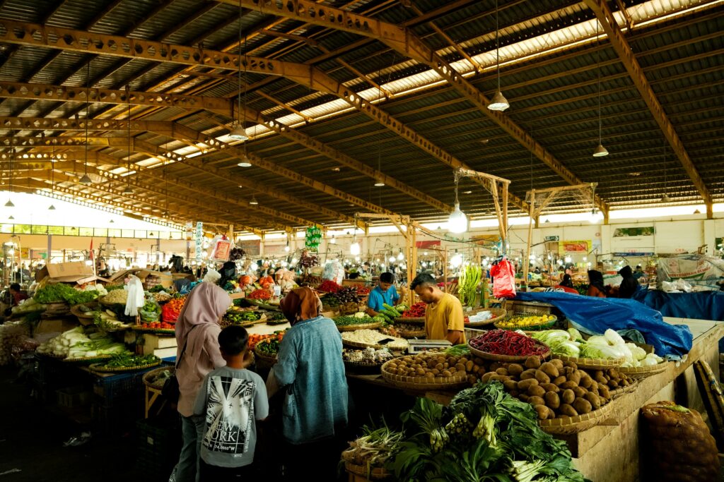 A bustling indoor market scene with diverse fresh fruits and vegetables on display under wooden roof.