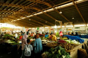 A bustling indoor market scene with diverse fresh fruits and vegetables on display under wooden roof.